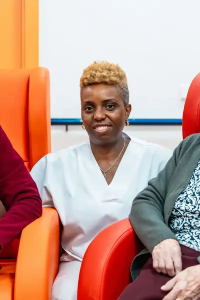 Verpleegster samen met twee bejaarde vrouwen, de bejaarde vrouwen zitten in stoelen en de verpleegster zit gehurkt tussen hen in.