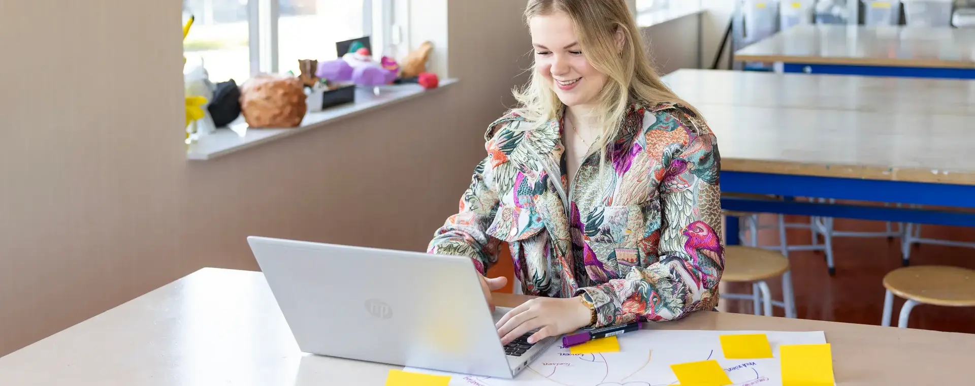 Student achter laptop in klaslokaal met notities en stiften