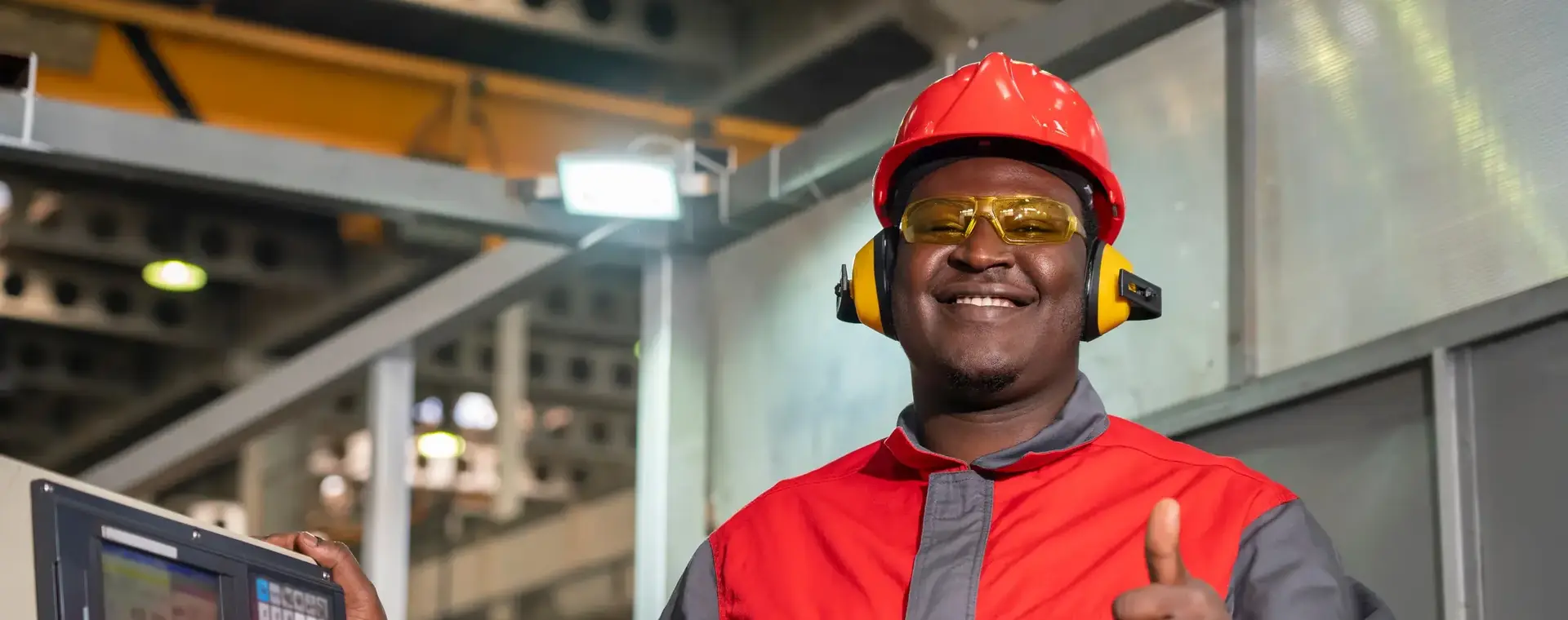 Student met ROCvA-overall, veiligheidsbril en oorbeschermers poseert naast machine in fabriek en steekt zijn duim op. 