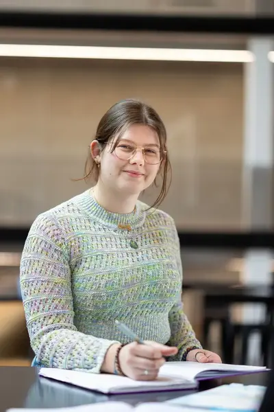 Student zit met een schrift aan tafel en kijkt glimlachend naar de camera.