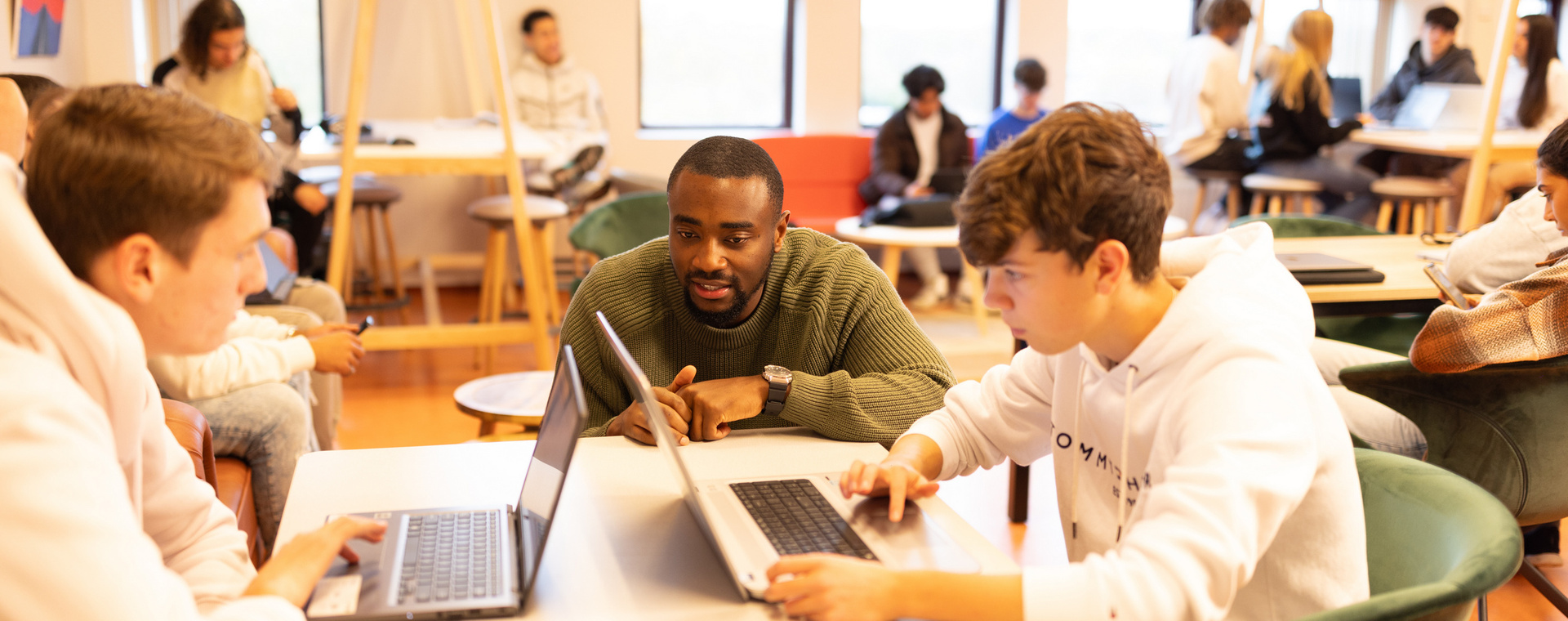 Docent begeleidt twee studenten tijdens het werken aan hun laptops in een lokaal met andere studenten op de achtergrond.