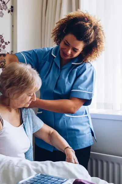 Een zorgmedewerker helpt een oudere vrouw bij het aankleden op bed. De sfeer is warm en zorgzaam.