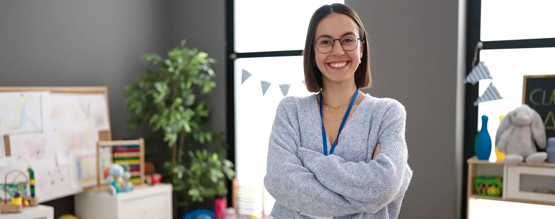 Een jonge vrouw staat lachend met haar armen over elkaar in een kleurrijke klasruimte. Op de achtergrond zijn speelgoed, knuffels en kindertekeningen te zien.