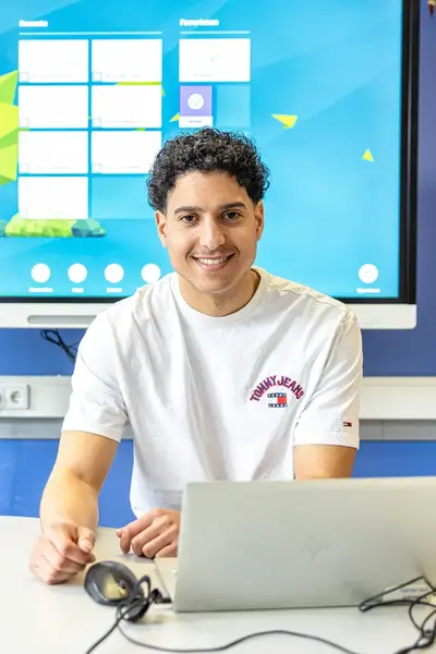 Docent zit aan een tafel met een laptop voor een digitaal schoolbord en kijkt lachend in de camera.