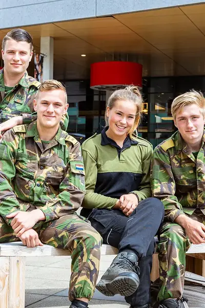 Vier studenten in legeruniform zitten en poseren lachend op houten banken buiten.