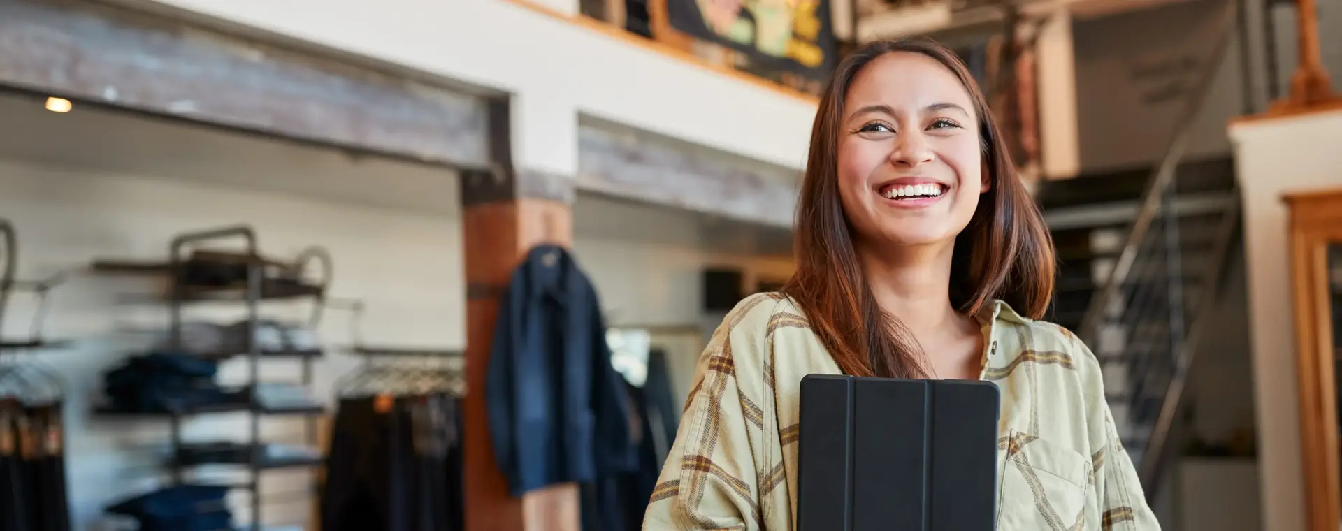 Een lachende medewerkster staat in een kledingwinkel. Ze heeft lang bruin haar en een ipad in haar arm.