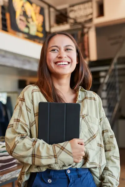 Een lachende medewerkster staat in een kledingwinkel. Ze heeft lang bruin haar en een ipad in haar arm.