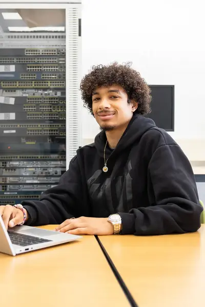 Student werkt op een laptop in een ICT-lokaal met netwerkapparatuur op de achtergrond. Hij glimlacht naar de camera terwijl hij aan tafel zit.        Ask ChatGPT