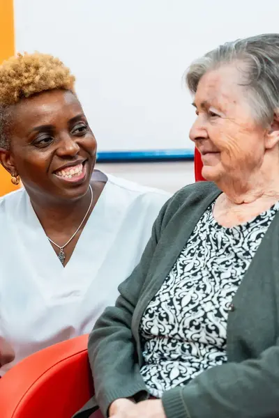 Gehurkte vrouw omarmt bejaarde dame in stoel