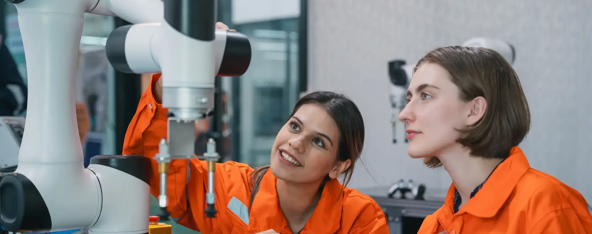 Twee vrouwen in oranje overalls bestuderen een robotarm