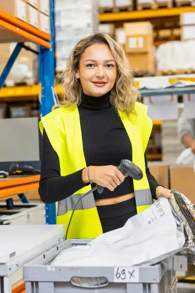 Een student werkt in een logistiek magazijn en scant een pakket met een barcodescanner. Ze draagt een gele veiligheidshes en staat bij een inpakstation.