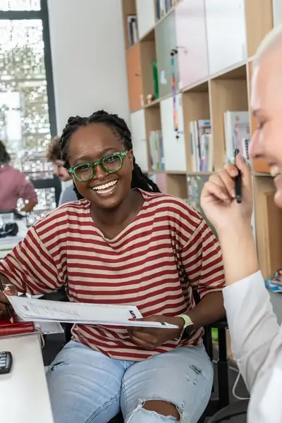 Twee vrouwen zitten samen aan een bureau in een klaslokaal of kantoorruimte. Ze lachen en overleggen terwijl één van hen een papier vasthoudt.