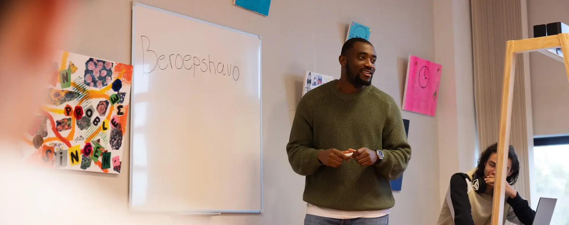 Een docent staat voor de klas bij een whiteboard met het woord 'Beroepshavo'. Hij spreekt de leerlingen aan, terwijl een student op de voorgrond aandachtig luistert.